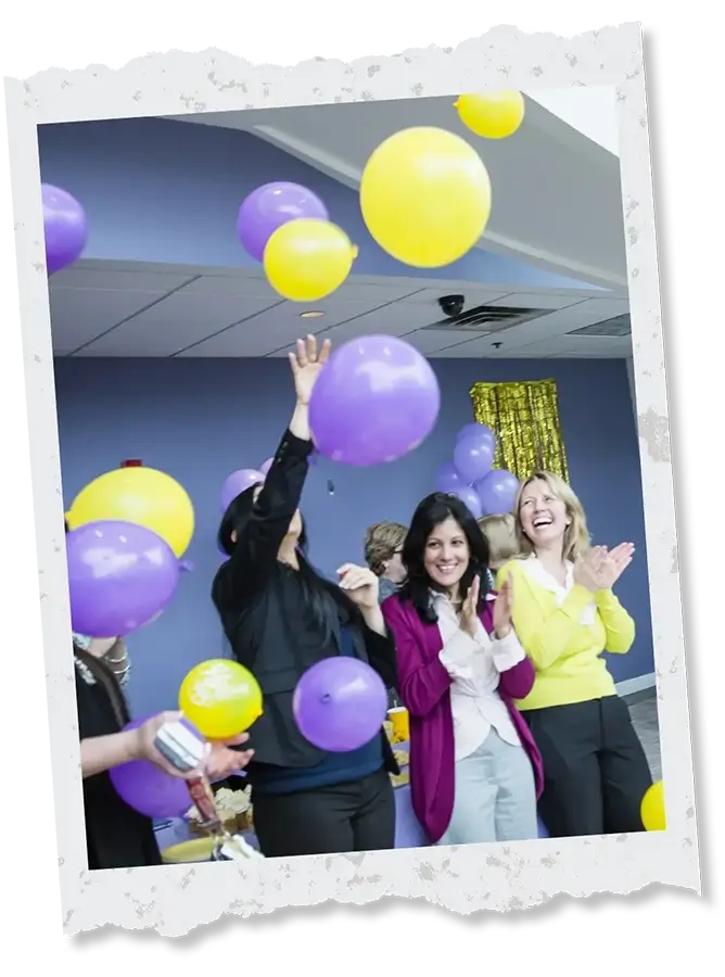 Three professional Women clapping and releasing purple and yellow balloons inside of a building