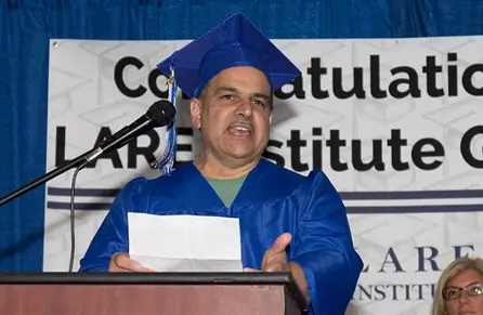 Adult male student wearing a blue cap and gown speaking at the podium during a graduation ceremony
